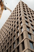 A tall, modern building with a grid-like facade made of beige bricks and large windows extends upwards. A yellow construction crane is visible adjacent to the building, suggesting ongoing construction or development work.