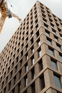 A tall, modern building with a grid-like facade made of beige bricks and large windows extends upwards. A yellow construction crane is visible adjacent to the building, suggesting ongoing construction or development work.
