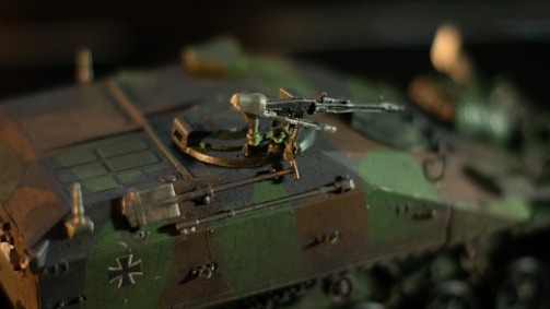 A detailed model of a military vehicle with camouflage patterns. A mounted machine gun is prominently displayed at the top, and a distinctive emblem is visible on the side. The lighting highlights the intricate design and textures of the model.