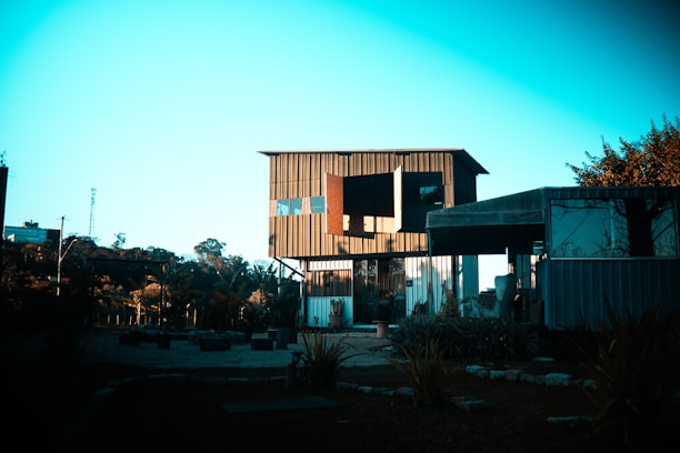 A modern building with a mix of glass and metal materials is situated in a serene environment with trees and foliage around. The structure features an open design with large windows and visible support beams. The sky is clear, allowing for bright natural light.