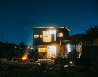 A modern two-story house with bright lights illuminating the front, set against a dark night sky filled with stars. A warm fire pit glows in the foreground, surrounded by seating arrangements. The scene is enriched by various plants and trees, providing a sense of tranquility.