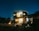 A modern two-story house with bright lights illuminating the front, set against a dark night sky filled with stars. A warm fire pit glows in the foreground, surrounded by seating arrangements. The scene is enriched by various plants and trees, providing a sense of tranquility.