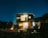 A modern two-story house with bright lights illuminating the front, set against a dark night sky filled with stars. A warm fire pit glows in the foreground, surrounded by seating arrangements. The scene is enriched by various plants and trees, providing a sense of tranquility.