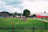 A rugby field surrounded by stands and a few structures. A group of players forms a huddle on the grass, and the weather appears to be overcast. The scene includes goalposts, a partially filled grandstand, and a large domed building in the background.