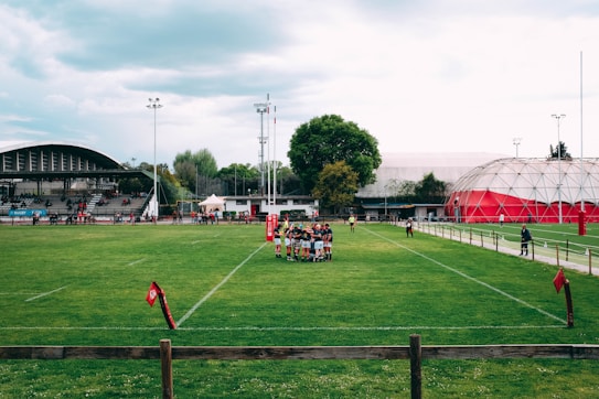 A rugby field surrounded by stands and a few structures. A group of players forms a huddle on the grass, and the weather appears to be overcast. The scene includes goalposts, a partially filled grandstand, and a large domed building in the background.