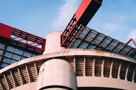 A modern architectural structure featuring large, curved concrete elements and striking red metal components. The design includes a combination of geometric patterns and industrial aesthetics, with glass panels allowing natural light to filter through.