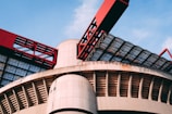 A modern architectural structure featuring large, curved concrete elements and striking red metal components. The design includes a combination of geometric patterns and industrial aesthetics, with glass panels allowing natural light to filter through.