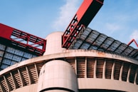 A modern architectural structure featuring large, curved concrete elements and striking red metal components. The design includes a combination of geometric patterns and industrial aesthetics, with glass panels allowing natural light to filter through.