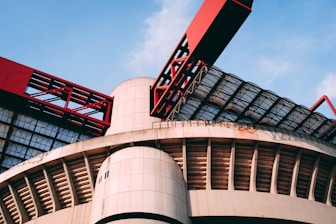 A modern architectural structure featuring large, curved concrete elements and striking red metal components. The design includes a combination of geometric patterns and industrial aesthetics, with glass panels allowing natural light to filter through.