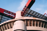 A modern architectural structure featuring large, curved concrete elements and striking red metal components. The design includes a combination of geometric patterns and industrial aesthetics, with glass panels allowing natural light to filter through.