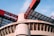 A modern architectural structure featuring large, curved concrete elements and striking red metal components. The design includes a combination of geometric patterns and industrial aesthetics, with glass panels allowing natural light to filter through.