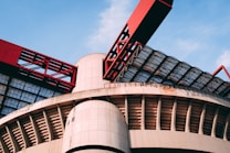 A modern architectural structure featuring large, curved concrete elements and striking red metal components. The design includes a combination of geometric patterns and industrial aesthetics, with glass panels allowing natural light to filter through.