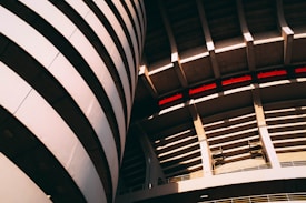 Curved architecture with large, sweeping lines that create an industrial, modern feel. The structure features arched elements and shadow play, highlighted by light and dark contrasts, with hints of red accents visible within the design.