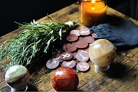 A table arrangement featuring a bundle of herbs, a lit amber candle, a black pouch, round wooden tokens with runic symbols, and three decorative gemstone spheres.