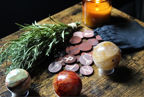 A table arrangement featuring a bundle of herbs, a lit amber candle, a black pouch, round wooden tokens with runic symbols, and three decorative gemstone spheres.