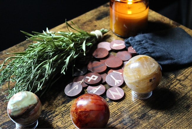 A table arrangement featuring a bundle of herbs, a lit amber candle, a black pouch, round wooden tokens with runic symbols, and three decorative gemstone spheres.