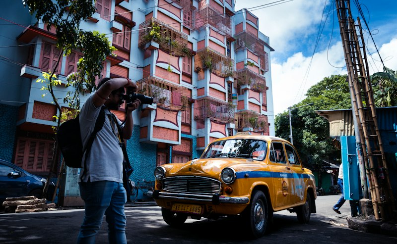 A yellow Ambassador taxi parked on a Kolkata street below a pink and pale blue apartment block with plants on its balcony grilles, a man crouching to photograph it