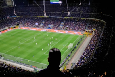 A large football stadium filled with spectators, where a match is taking place on a well-lit green field. The scene is viewed from a high vantage point with a silhouette of a person in the foreground.