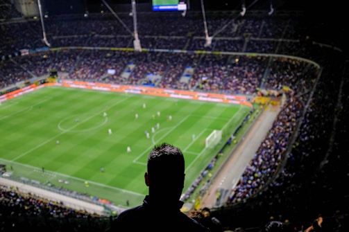 A large football stadium filled with spectators, where a match is taking place on a well-lit green field. The scene is viewed from a high vantage point with a silhouette of a person in the foreground.
