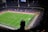 A large football stadium filled with spectators, where a match is taking place on a well-lit green field. The scene is viewed from a high vantage point with a silhouette of a person in the foreground.