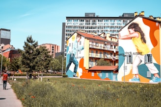 A vibrant urban scene with large, colorful murals painted on the sides of two buildings. One mural depicts a person pushing against a wall with the phrase 'Close the Gap' while the other shows a person with the text 'Open Your Future'. The murals are set against a backdrop of residential buildings and a clear blue sky. In the foreground, a grassy area is visible with blooming flowers and a sidewalk where a person is walking away from the camera.