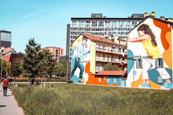 A vibrant urban scene with large, colorful murals painted on the sides of two buildings. One mural depicts a person pushing against a wall with the phrase 'Close the Gap' while the other shows a person with the text 'Open Your Future'. The murals are set against a backdrop of residential buildings and a clear blue sky. In the foreground, a grassy area is visible with blooming flowers and a sidewalk where a person is walking away from the camera.