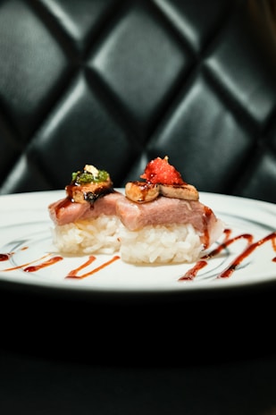 Two pieces of sushi are elegantly arranged on a white plate, garnished with vibrant toppings like green herbs and red roe. The plate is artistically accented with lines of red sauce. The background is a tufted, dark leather surface, giving a sophisticated ambiance.