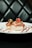 Two pieces of sushi are elegantly arranged on a white plate, garnished with vibrant toppings like green herbs and red roe. The plate is artistically accented with lines of red sauce. The background is a tufted, dark leather surface, giving a sophisticated ambiance.
