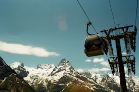 A gondola lift is suspended in the air against a backdrop of snow-covered mountains and a clear blue sky. A metallic cable tower supports the lift, which is transporting passengers. The majestic peaks are partially covered with snow, and a fluffy white cloud floats in the sky.