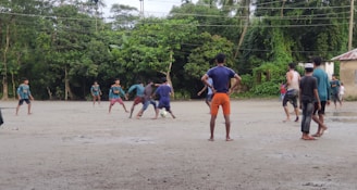 A group of people are playing football on a dirt field surrounded by lush green trees. The players are in casual clothing, with one kicking a ball while others look on. The sky is overcast, suggesting a recent or impending rain.