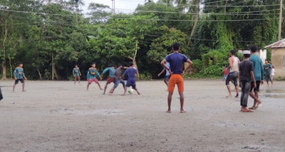 A group of people are playing football on a dirt field surrounded by lush green trees. The players are in casual clothing, with one kicking a ball while others look on. The sky is overcast, suggesting a recent or impending rain.