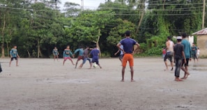 A group of people are playing football on a dirt field surrounded by lush green trees. The players are in casual clothing, with one kicking a ball while others look on. The sky is overcast, suggesting a recent or impending rain.