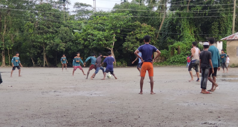 A group of people are playing football on a dirt field surrounded by lush green trees. The players are in casual clothing, with one kicking a ball while others look on. The sky is overcast, suggesting a recent or impending rain.