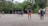 A group of people are playing football on a dirt field surrounded by lush green trees. The players are in casual clothing, with one kicking a ball while others look on. The sky is overcast, suggesting a recent or impending rain.
