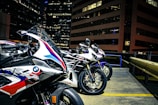 Three sport motorcycles are parked in an urban setting, likely on a rooftop with tall office buildings in the background. The motorcycles feature sleek designs and branding from manufacturers, indicating they are high-performance models. The environment is illuminated by city lights, revealing details of the bikes' paint jobs and textures.