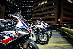 Three sport motorcycles are parked in an urban setting, likely on a rooftop with tall office buildings in the background. The motorcycles feature sleek designs and branding from manufacturers, indicating they are high-performance models. The environment is illuminated by city lights, revealing details of the bikes' paint jobs and textures.