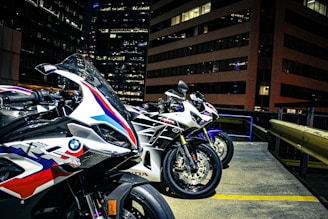 Three sport motorcycles are parked in an urban setting, likely on a rooftop with tall office buildings in the background. The motorcycles feature sleek designs and branding from manufacturers, indicating they are high-performance models. The environment is illuminated by city lights, revealing details of the bikes' paint jobs and textures.