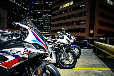 Three sport motorcycles are parked in an urban setting, likely on a rooftop with tall office buildings in the background. The motorcycles feature sleek designs and branding from manufacturers, indicating they are high-performance models. The environment is illuminated by city lights, revealing details of the bikes' paint jobs and textures.