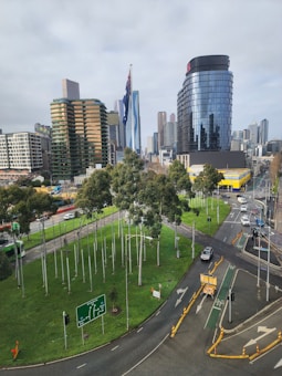 A cityscape featuring a large intersection with green traffic signs and directional road markings. Tall modern buildings with glass facades line the skyline, and there are trees and green spaces near the roads. Vehicles including cars and a tram are visible on the streets.