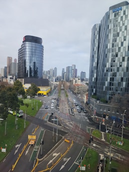 The image depicts a modern urban street scene with high-rise buildings, including a shiny black cylindrical tower and a distinctive multi-faceted building. The street below features multiple lanes, traffic lights, and pedestrian signals. There are green spaces with trees and a yellow building in the background.