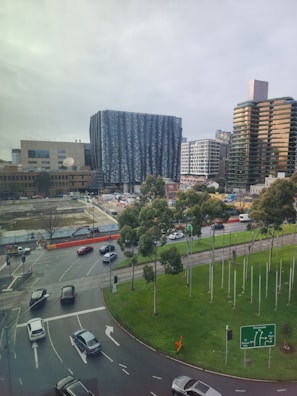 A cityscape displaying modern high-rise buildings and a prominent architecturally styled structure with a blue and white patterned facade. Below, a busy urban street intersection shows vehicles navigating multiple lanes. Trees line the road near the intersection, and a construction site occupies a section of the open space.