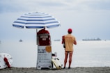 Two lifeguards are at a beach, one sitting on a lifeguard chair under a blue and white striped umbrella and the other standing nearby. They are dressed in lifeguard attire with the word 'Lifeguard' printed on their shirts. In the background, ships can be seen on the water.