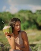 A woman with dark hair is sitting in a natural setting, holding a fresh coconut with a straw near her face. She is wearing a sleeveless knit top and appears to be relaxed, gazing into the distance. The background is a blurry green landscape, suggesting an outdoor or tropical environment.