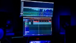 A dimly lit room features two large computer screens displaying complex audio editing software. Multiple speakers are visible in the background, with a digital clock and other electronic equipment situated below the screens.