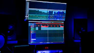 A dimly lit room features two large computer screens displaying complex audio editing software. Multiple speakers are visible in the background, with a digital clock and other electronic equipment situated below the screens.