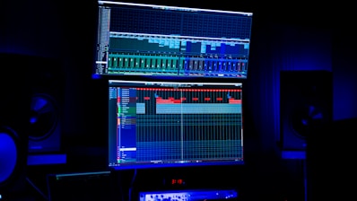 A dimly lit room features two large computer screens displaying complex audio editing software. Multiple speakers are visible in the background, with a digital clock and other electronic equipment situated below the screens.