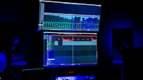 A dimly lit room features two large computer screens displaying complex audio editing software. Multiple speakers are visible in the background, with a digital clock and other electronic equipment situated below the screens.