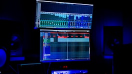 A dimly lit room features two large computer screens displaying complex audio editing software. Multiple speakers are visible in the background, with a digital clock and other electronic equipment situated below the screens.