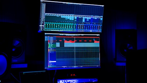 A dimly lit room features two large computer screens displaying complex audio editing software. Multiple speakers are visible in the background, with a digital clock and other electronic equipment situated below the screens.