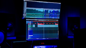 A dimly lit room features two large computer screens displaying complex audio editing software. Multiple speakers are visible in the background, with a digital clock and other electronic equipment situated below the screens.